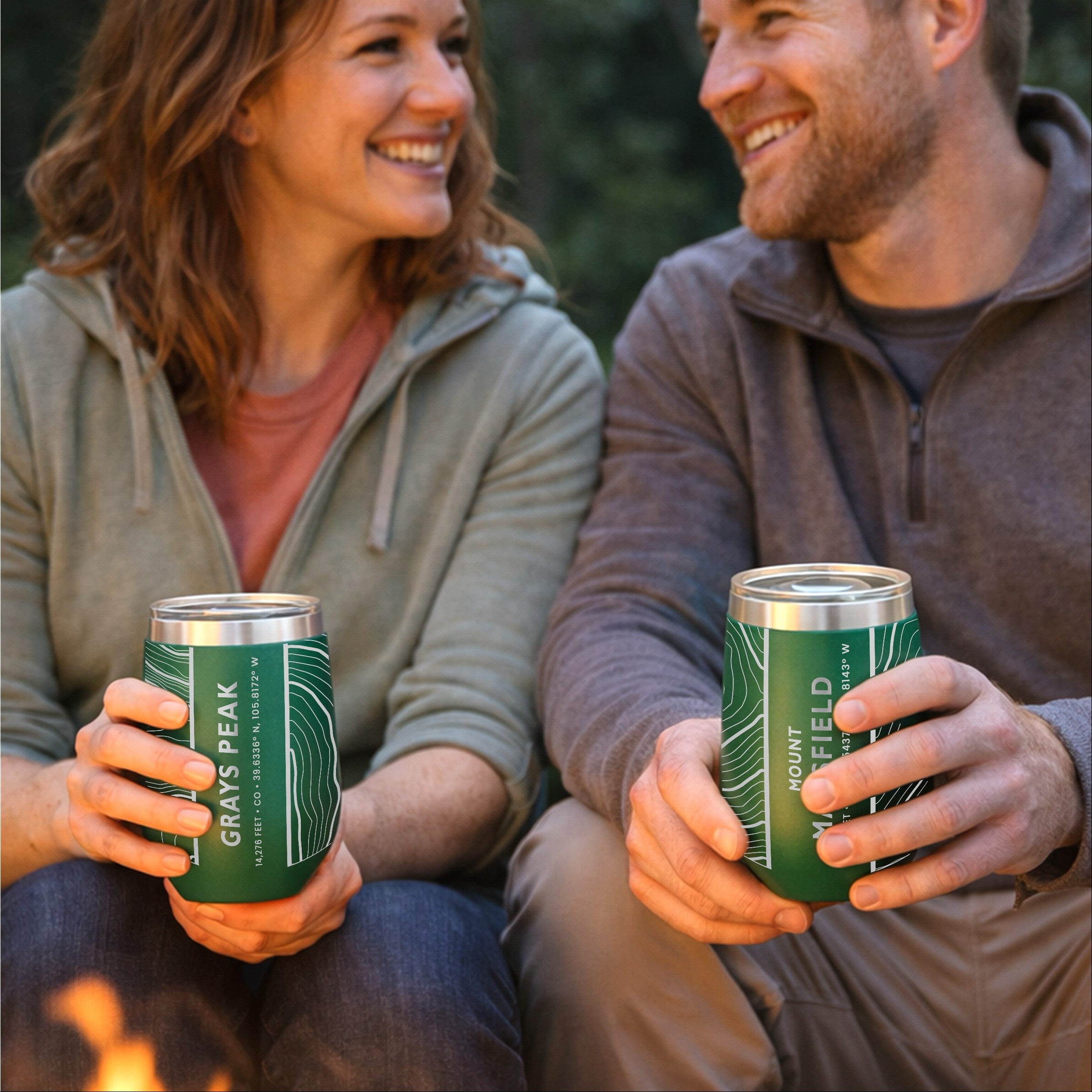 A couple sitting by a campfire holding green wine tumblers featuring topographic map designs of Grays Peak and Mount Mansfield.