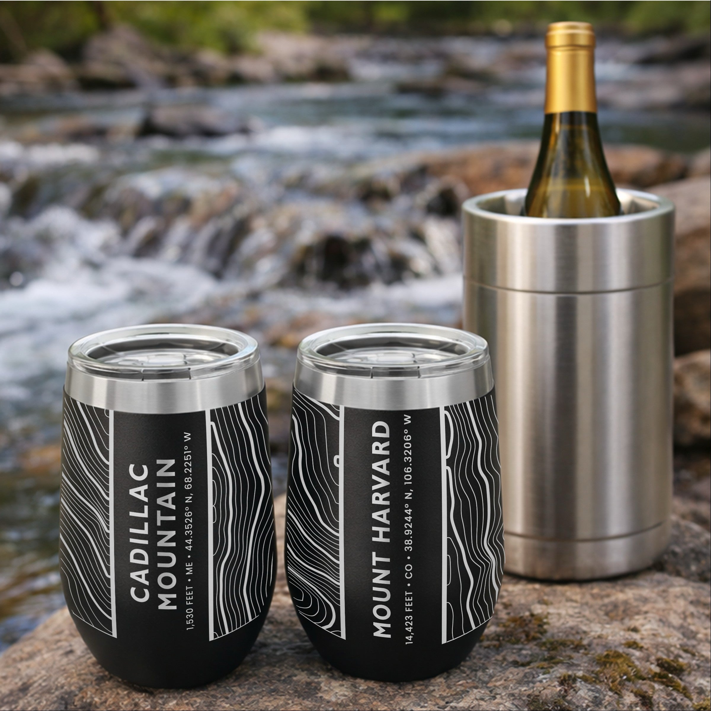 Two wine tumblers on a rock by a river with a wine bottle in a cooler near by.  Tumblers feature topo maps of Cadilac Mountain and Mount Harvard.